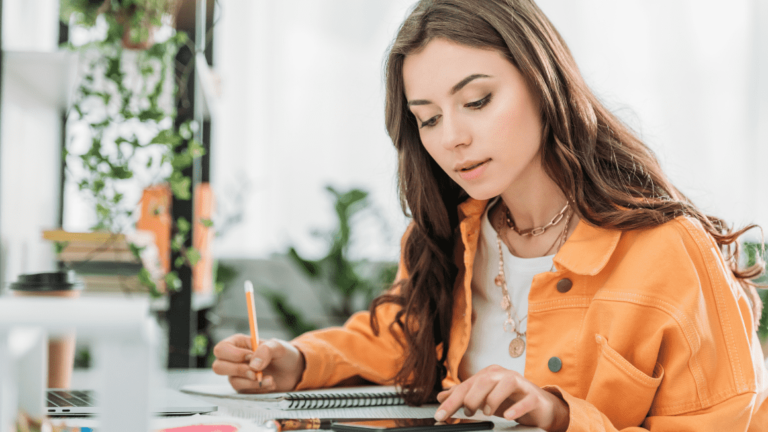 Woman sitting at desk using smartphone and writing in notebook