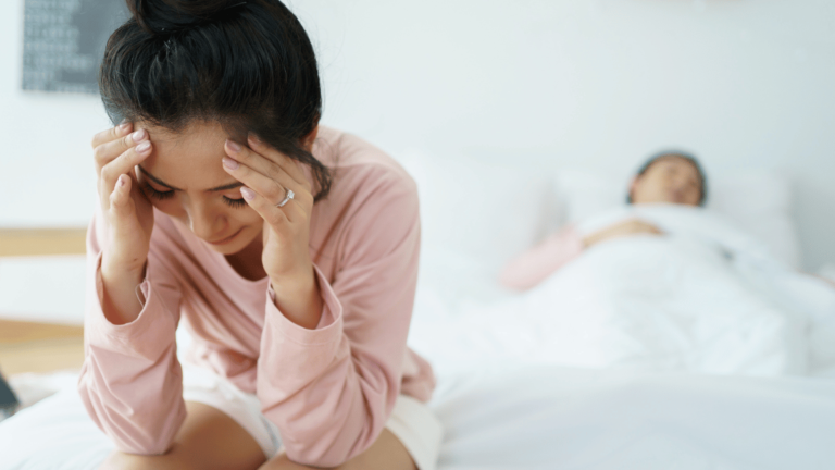 upset Asian woman sitting and crying on the bed while her husband is sleeping