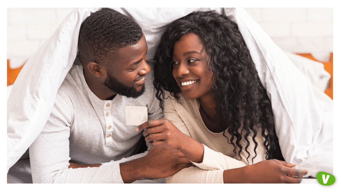 african american couple holding condom, lying on bed under white blanket, looking at each other with love and care. safe sex concept