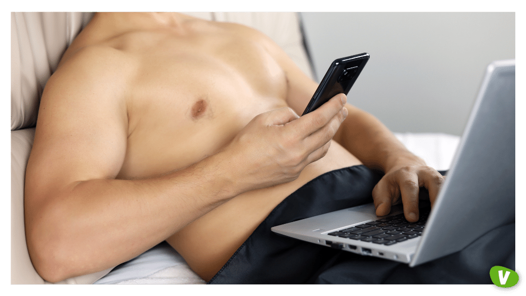 muscular man lying in bed under a black blanket with laptop and smartphone in hands