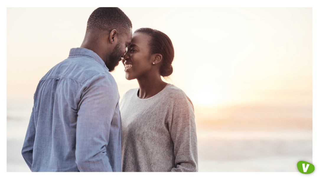 smiling young African couple talking and laughing together while standing face to face on a beach at sunset