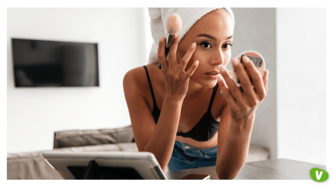 close up portrait of a young asian woman with bath towel on head doing her make up while leaning on a table and looking at the mirror