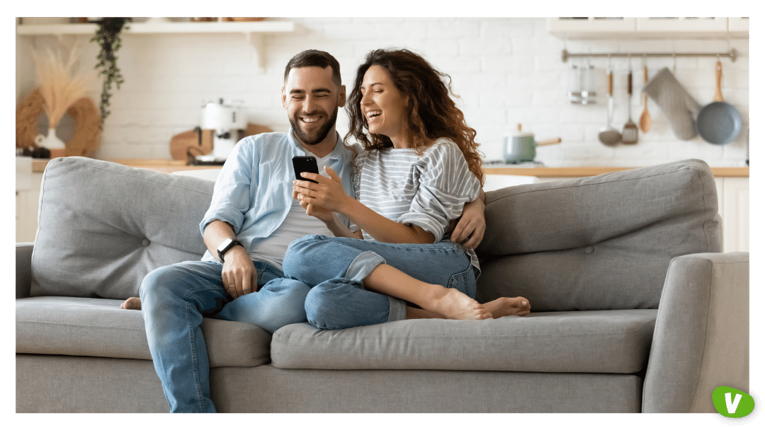 happy young woman and man hugging, using smartphone together, sitting on cozy couch at home