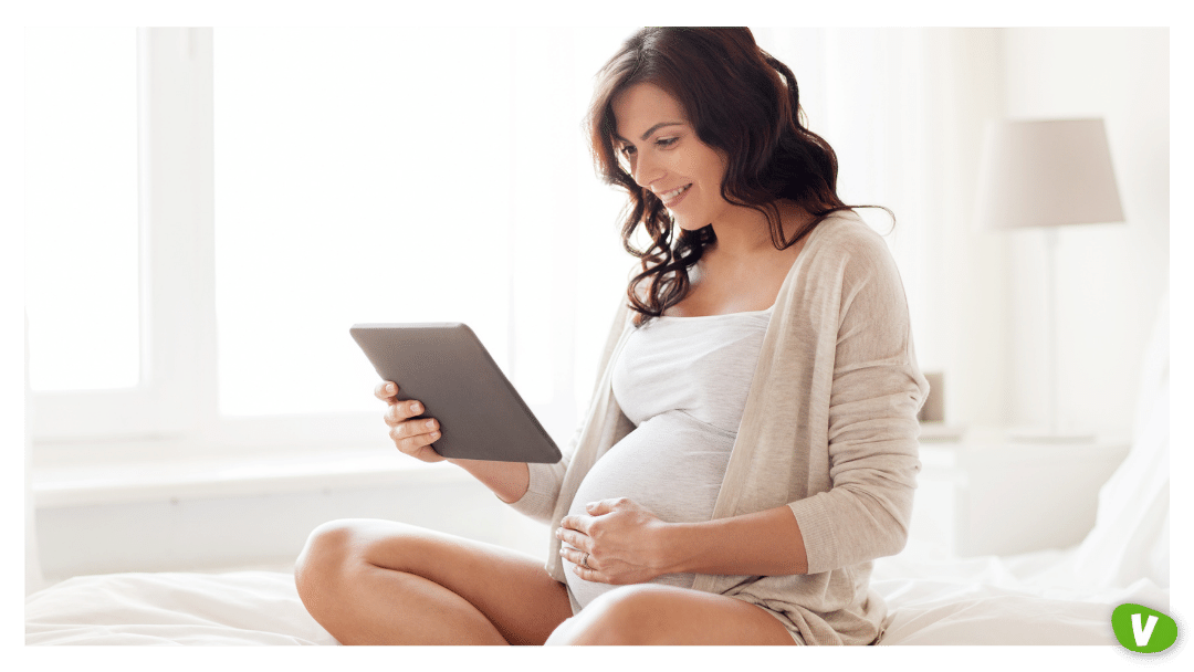 a pregnant woman holding her belly while looking into a tablet on her bed