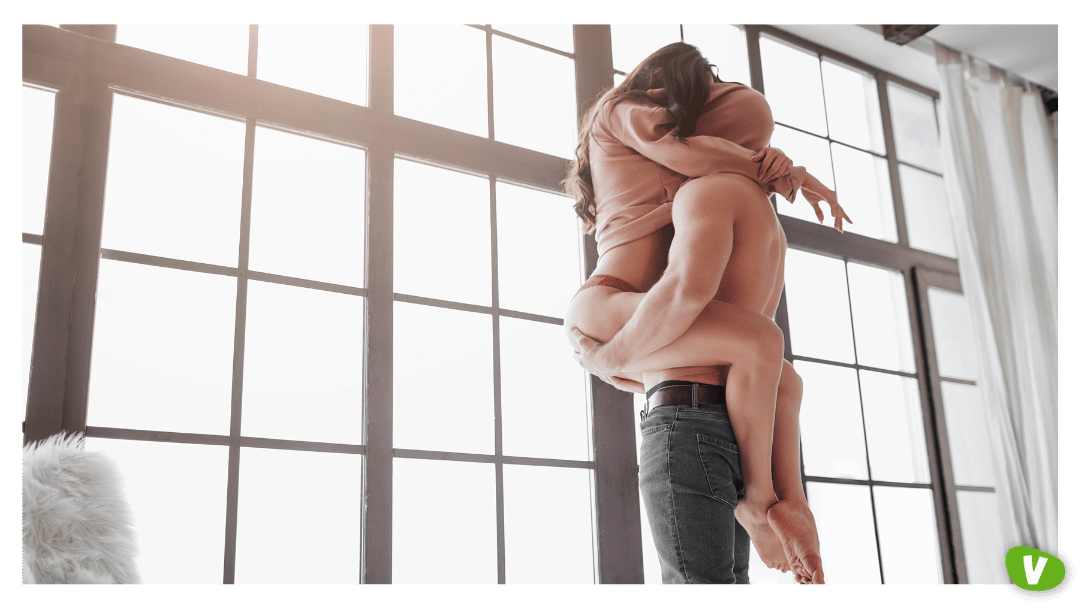 Sexy Couple Stand at Window and Hide Head under Sweater. Guy Hold Woman on Hands. She Sit on Him and Embrace His Body with Legs.