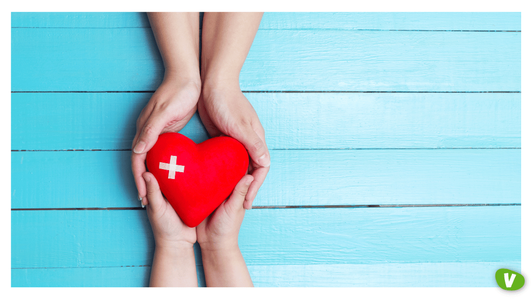 Two pairs of hands gently holding a red heart with a white medical cross, symbolising care, health support, and protection.