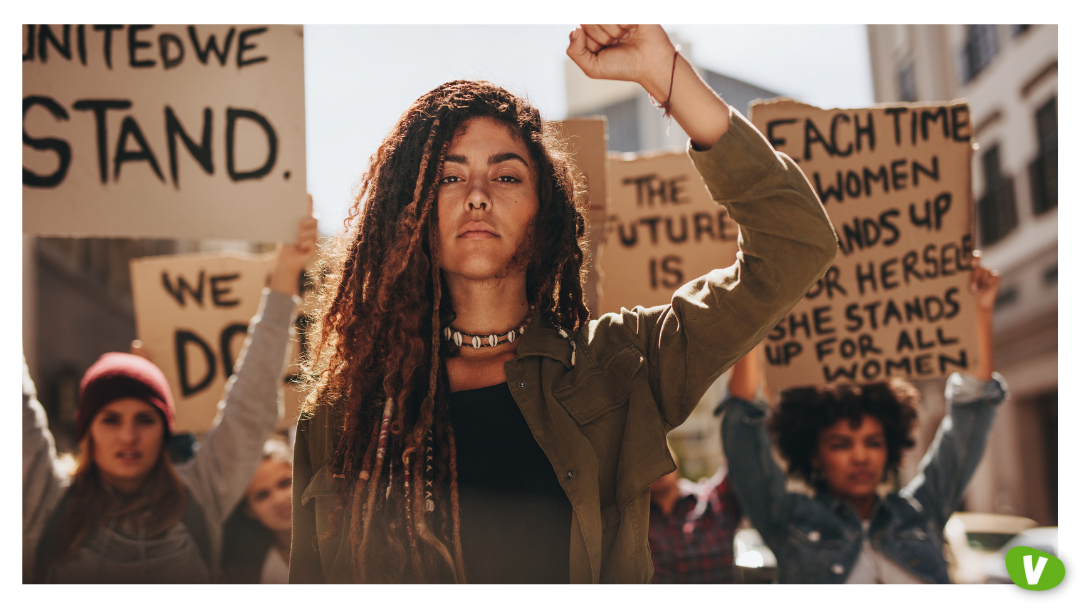 A group of women protesting with empowering signs, including ‘United We Stand’ and ‘She Stands Up for All Women’, led by a confident woman raising her fist. Concept of feminism and sex work.