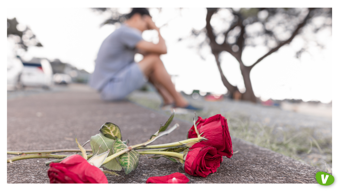Red roses lying on the pavement in the foreground with a man sitting blurred in the background, head in hands, portraying heartbreak and rejection often felt when catching feelings for an escort.