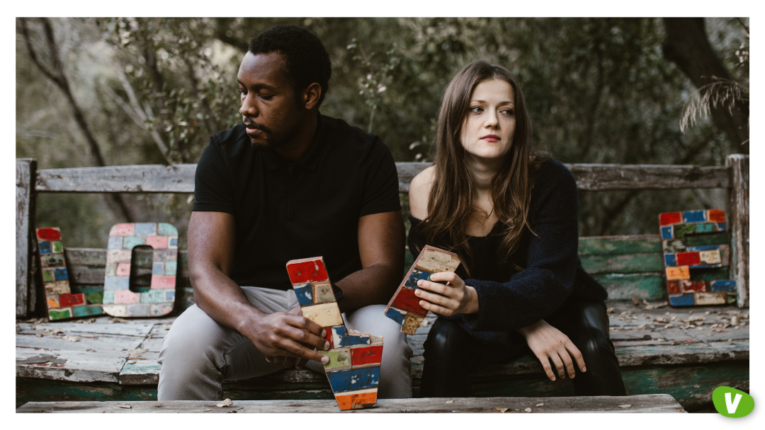 A couple sitting apart on a weathered bench, both looking away with serious expressions, holding mismatched wooden letters, symbolizing a fractured relationship — representing the emotional struggles of catching feelings for an escort.