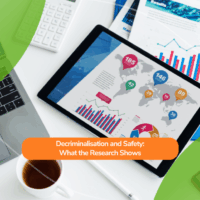 A workspace with a laptop, tablet, documents, and a cup of tea. The tablet displays colourful data charts and maps, representing research findings. Overlaid text reads: “Decriminalisation and Safety: What the Research Shows.”