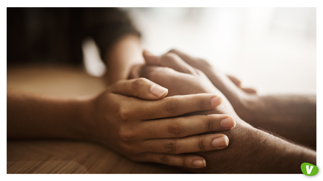 Two people hold hands gently across a table. One person’s hands rest comfortingly on top of the other’s, symbolising care, support, and reassurance.
