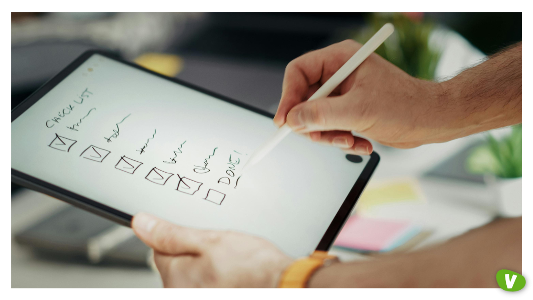 A person holds a digital tablet and uses a stylus to check off items on a handwritten checklist. Several boxes are ticked, and the final item is marked “DONE!” The background is a blurred workspace.