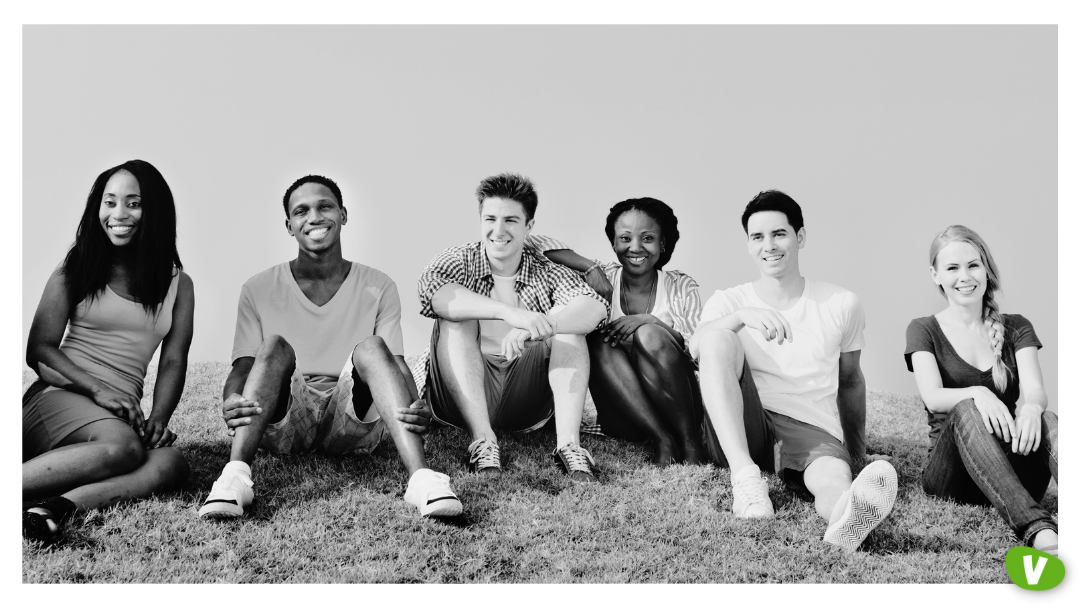 Six young adults sitting in a row on grass outdoors, smiling and relaxed, facing the camera in a casual group portrait.