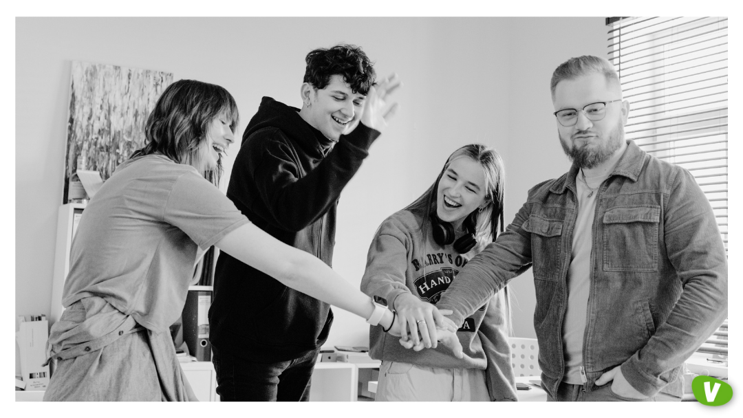 A group of four young adults standing together indoors, smiling and placing their hands in a stack as a gesture of teamwork and unity.