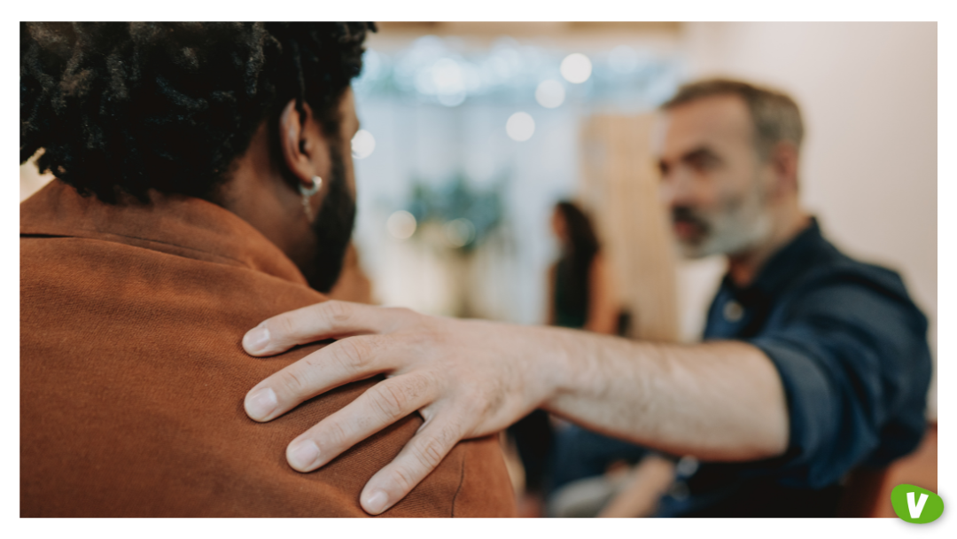 A person resting their hand on another person’s shoulder in a gesture of comfort during a conversation in a softly lit indoor setting.
